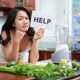 Pretty Asian woman frowning and holding paper saying Help standing with heap of green vegetables and blender in kitchen